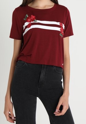 Vrouw draagt een kort mouw crop top in bordeauxrood met witte strepen en borduurwerk van een rode roos, gecombineerd met een zwarte high-waisted jeans.