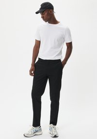 Camiseta blanca de manga corta, pantalones negros ajustados y zapatillas de correr plateadas con detalles en azul. Gorra negra con una etiqueta. Postura de pie.