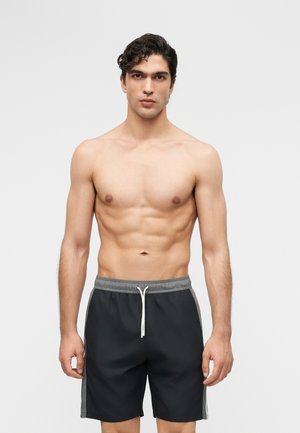 Młody mężczyzna o ciemnych kręconych włosach, bez koszulki, w czarno-szarych kąpielówkach z białym troczkiem, stojący na tle jasnego, jednolitego tła.