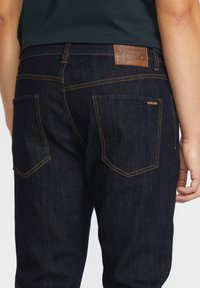Jean bleu foncé avec une coupe fuselée, fabriqué en denim. Présente des coutures orange, une étiquette en cuir à l'arrière et des poches arrière standard.