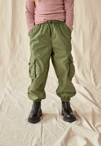 Grüne Cargo-Hose mit elastischen Bündchen und Taschen, kombiniert mit schwarzen Knöchelstiefeln, vor einem hellen Stoffhintergrund.