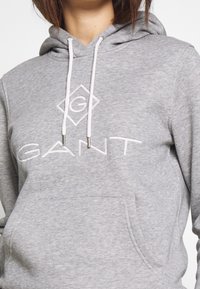 Primo piano di una persona che indossa un felpa leggera grigia con cappuccio, con lacci bianchi e il logo "GANT" ricamato sul petto.