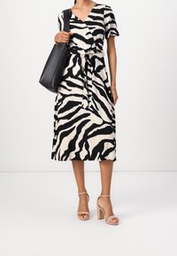 Šaty s černobílým zebrovaným vzorem, s krátkým rukávem, páskem v pase a midi délkou, doplněné o nudové sandály na blokovém podpatku a černou tašku.