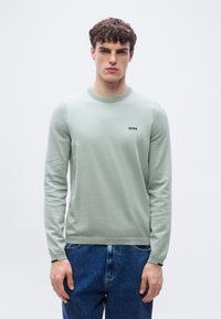 Νέος με κοντά σγουρά μαλλιά, φορώντας μακριά πράσινη μπλούζα με το λογότυπο "BOSS" και μπλε τζιν, στέκεται μπροστά σε απλό φόντο.