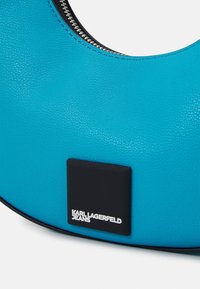 Turkos läderhandväska med rundad form, utrustad med en silverfärgad dragkedja och en svart fyrkantig detalj med "KARL LAGERFELD JEANS" i vitt.