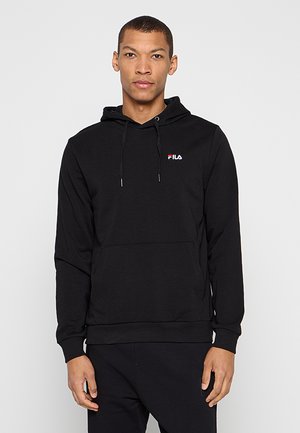 Homme portant un sweat à capuche noir uni FILA avec poche kangourou, debout devant un fond gris clair.
