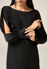 Czarna sukienka z wycięciami na rękawach i gładką fakturą. Długie rękawy z marszczonymi mankietami. Wyraziste kolczyki z kolorowymi kamieniami szlachetnymi.