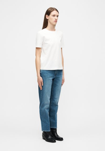 Νεαρή γυναίκα που στέκεται και κοιτάζει ελαφρώς προς τα δεξιά, φοράει λευκό πουκάμισο με κοντά μανίκια, μπλε τζιν και μαύρα μποτάκια αστραγάλου σε λευκό φόντο.