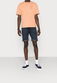 Camiseta corta de manga en color durazno claro con un pequeño gráfico, combinada con pantalones cortos de mezclilla oscuros remangados en los puños y zapatillas navy con suelas blancas.