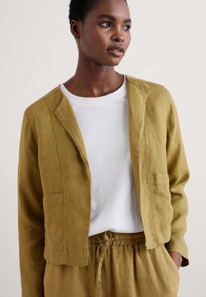 Helle Senf-Linnenblazer mit einem lockeren Schnitt, leicht verkürztem Design, Reverskragen und zwei vorderen Taschen, getragen über einem weißen Hemd.