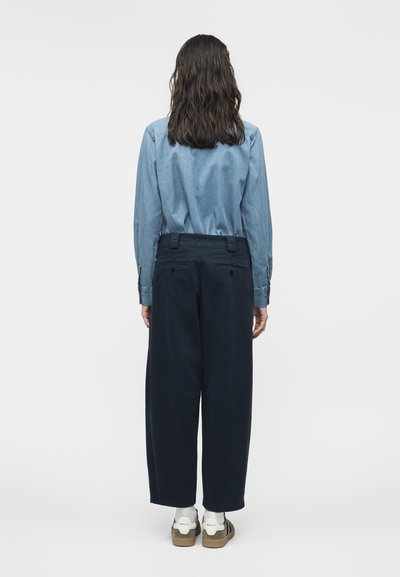 Άτομο με μακριά σκούρα μαλλιά, ντυμένο με μπλε μακρυμάνικο πουκάμισο, σκούρο φαρδύ παντελόνι και λευκά αθλητικά παπούτσια, στέκεται γυρισμένο προς τα πίσω.