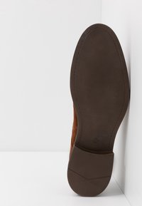 Chaussure en daim marron avec une semelle en caoutchouc texturée, présentant une forme lisse et incurvée et un bout arrondi, positionnée sur un fond blanc.