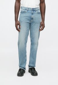 Gaiši zili džinsa džinsi ar taisnu piegriezumu, piecām kabatām, smalku izbalējumu un pogu aizdari, kopā ar melniem apaviem.