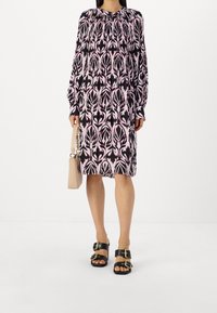 Šaty s dlouhým rukávem, které mají černý a růžový květinový vzor, kulatý výstřih a nařasený pas. Doplněné o černé sandály a béžovou tašku.