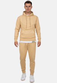 Beige hoodie met trekkoorden en een voorzak, gecombineerd met bijpassende joggers met elastische boorden, beide gemaakt van zachte stof. Witte sneakers.