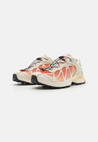 Des chaussures de course légères avec une tige en mesh beige et des accents orange, présentant un design ondulé unique, une semelle extérieure noire et un système de laçage élastique.