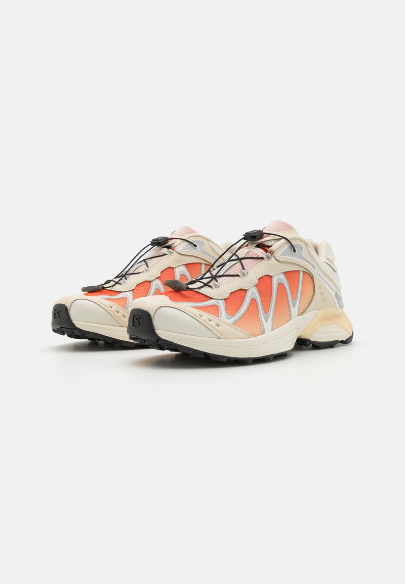 Zapatillas de running ligeras con una parte superior de malla beige y acentos naranjas, que presentan un diseño de ondas único, suela negra y un sistema de cordones elásticos.