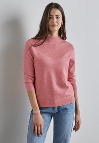 Różowy sweter dzianinowy z wysokim ściągaczem, z chevronowym wzorem na przodzie i ściągaczami na rękawach, w połączeniu z jasnoniebieskimi dżinsami.