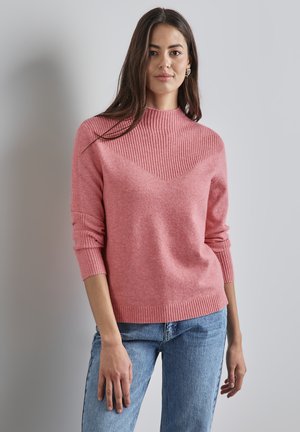 Gebreide roze trui met een hoge geribde hals, voorzien van een visgraatpatroon aan de voorkant en geribde manchetten, gecombineerd met lichtblauwe spijkerbroek.