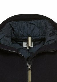 Zwarte fleecejack met een marineblauwe gevoerde capuchon, voorzien van een zilveren rits en een stoffen label met maat 38. Soepele interieur textuur.
