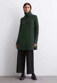 Túnica de malha verde escura com gola alta, apresentando um padrão texturado, mangas compridas e fendas laterais, combinada com calças pretas de perna larga.