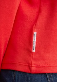 Rød bomuldstrøje med et mærke, der læser "Marc O'Polo DENIM" nær sømmen, med en glat tekstur og afslappet design.