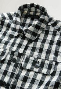 Cămașă în carouri cu un model alb-negru, material texturat, cu guler și două buzunare la piept cu nasturi. Prezintă nasturi negri.
