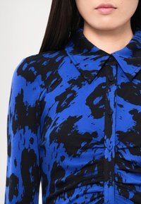 Kék-fekete mintás hosszú ujjú ing galléros nyakkivágással, gombos lezárással és a testen összegyűrt textúrával.