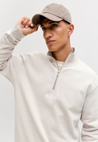 Jaunas vyras reguliuoja smėlio spalvos kepurę, dėvi šviesiai pilką ketvirčio užtrauktuko džemperį su logotipu, neutralaus fonas, žiūri šiek tiek į šoną.