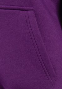 La tela morada presenta un bolsillo tipo canguro con una textura suave y un detalle de costura visible. El material parece blando y ligero.