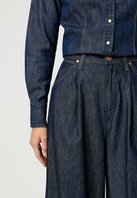 Camicia in denim scuro con bottoni a pressione, caratterizzata da un design aderente, e pantaloni larghi in denim scuro con cuciture visibili e tessuto testurizzato.