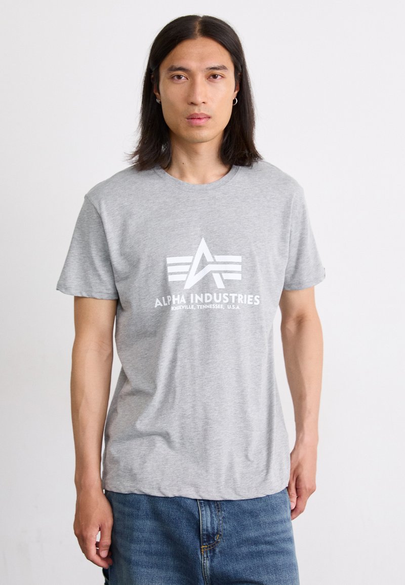 Pilkas medvilnės marškinėliai su balta atspausdinta "ALPHA INDUSTRIES" logotipu, turetu žvaigždės ir juostų dizainą. Trumpomis rankovėmis, laisvas tinkamas.