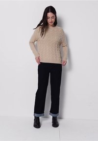 Beige Strickpullover mit Rundhalsausschnitt, kombiniert mit hochgeschnittenen schwarzen Jeans und Ankle Boots. Model steht vor einem schlichten Hintergrund.
