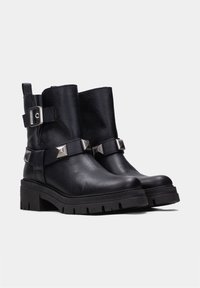 Botas de tobillo de cuero negro con un acabado texturizado, que cuentan con una correa ancha con tachuelas metálicas y un acento de hebilla. Diseño de suela gruesa y estriada.