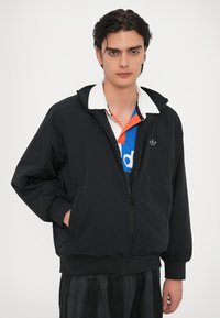 Crna jakna na zatvaranje s kapuljačom, s elastičnim rukavima i suptilnim logotipom na prsima. Nosila se preko višebojnog dolgava.