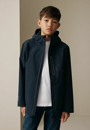 Dreng iført en marineblå hættetrøje over en hvid skjorte og blå jeans, stående mod en ensfarvet beige væg.