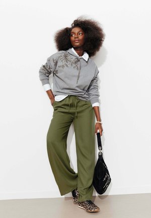 Femme aux cheveux bouclés et volumineux porte un sweat à capuche gris orné, une chemise blanche, un pantalon large olive, des chaussures à imprimé léopard, tenant un sac à main noir.