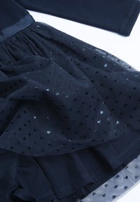 Námořnické šaty s přiléhavým topem a vrstvenou tylovou sukní. Tyl je zdobený černými vzory ve tvaru srdcí a má jemný, texturovaný povrch.