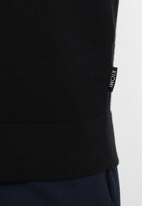 Pull noir en maille avec un ourlet côtelé et une petite étiquette KLOMI sur le côté. Le tissu semble doux et texturé, adapté pour un usage décontracté.