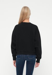 Svart bomullssweatshirt med cropped design, senkende skuldre og ribbede mansjetter. Kombinert med blå denimjeans. Vanlig bakfra.