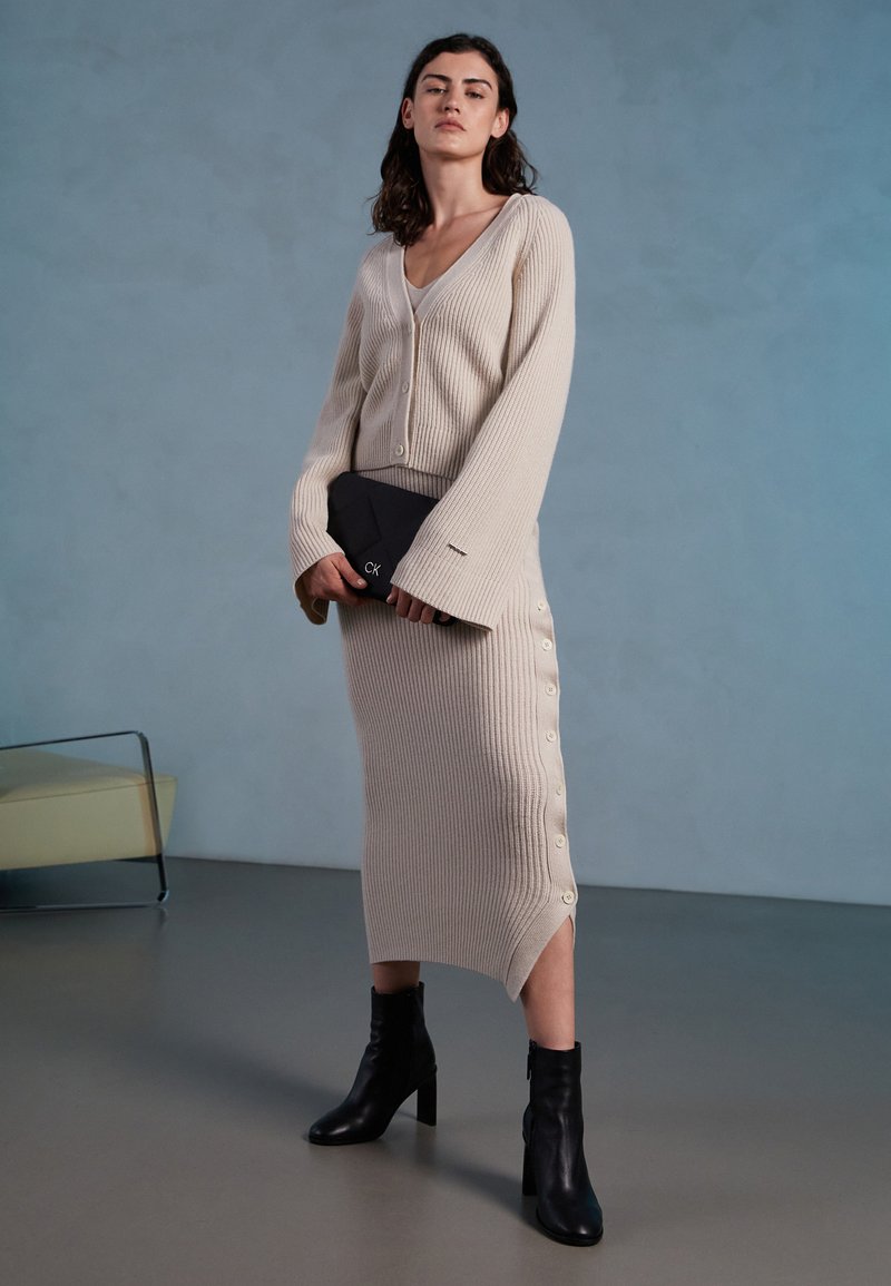 Vestido de punto acanalado beige con mangas largas y botones laterales; combinado con un bolso de mano negro y botas de tobillo. Diseño minimalista sobre un fondo neutro.