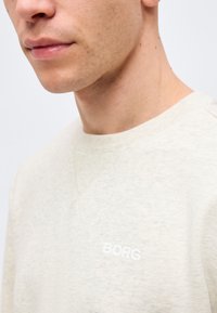 Κοντινό πλάνο ενός άνδρα που φοράει ανοιχτό μπεζ φούτερ με κεντημένο το λευκό γράμμα "BORG" στο στήθος.