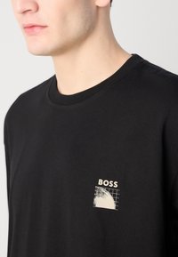 Černé bavlněné tričko s malou krémovou grafikou s mřížkovaným vzorem a textem "BOSS" na levé části hrudi. Design s kulatým výstřihem.