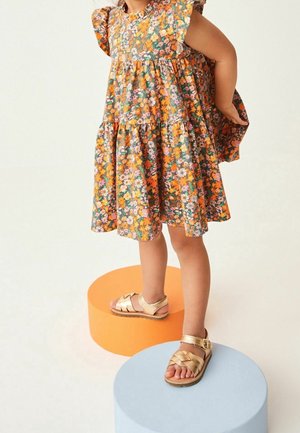 Robe à motifs floraux en orange, violet et vert ; manches à volants et design en étages. Porte des sandales dorées avec une boucle. Se tenant sur des piédestaux ronds.