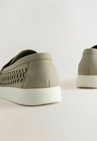 Lys grå læderslippers med perforerede detaljer, hvide såler og strukturerede overflader. Bagfra fremhæver designets unikke accenter.