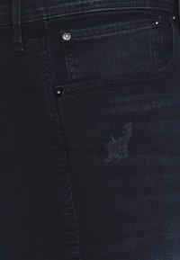 Tummansiniset farkut slim fit -leikkauksella, joissa on pieni kulunut alue edessä. Kankaan pinta on sileä ja se on korostettu metalliosilla.