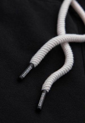 Cordon de capuche torsadé blanc avec embouts en plastique noir portant l'inscription "OMBRE" sur un fond en tissu noir.