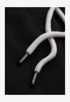 Cordon de capuche torsadé blanc avec embouts en plastique noir portant l'inscription "OMBRE" sur un fond en tissu noir.