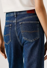 Temno modre kavbojke s klasičnim krojem. Imajo dva zadnja žepa z kontrastnim šivom in usnjen logotip na pasu.