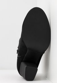 Zwarte enkellaars van suède met blokhak, getextureerde zool, ritssluiting aan de zijkant en een gladde afwerking. Het ontwerp is eenvoudig en functioneel.